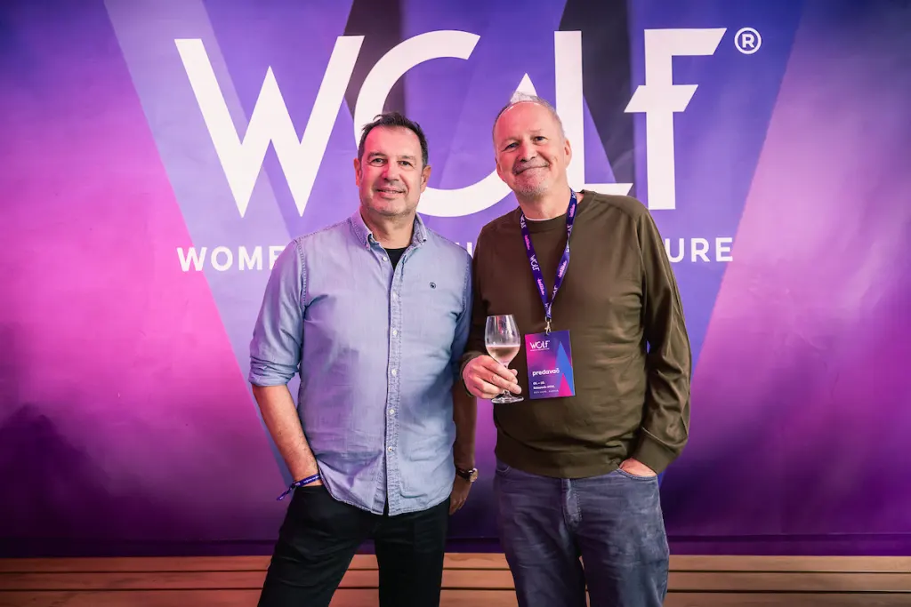 Two individuals pose together at a WCWF event, one holding a wine glass, against a purple backdrop featuring the event's logo.