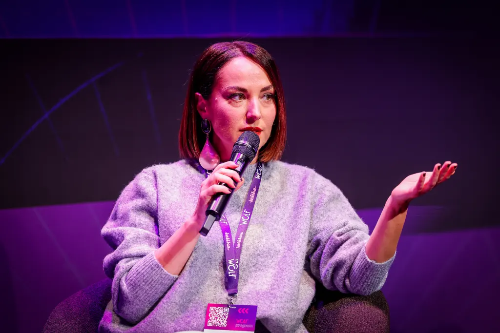 A person speaks into a microphone while seated on a stage bathed in purple lighting, wearing a lanyard with event branding.