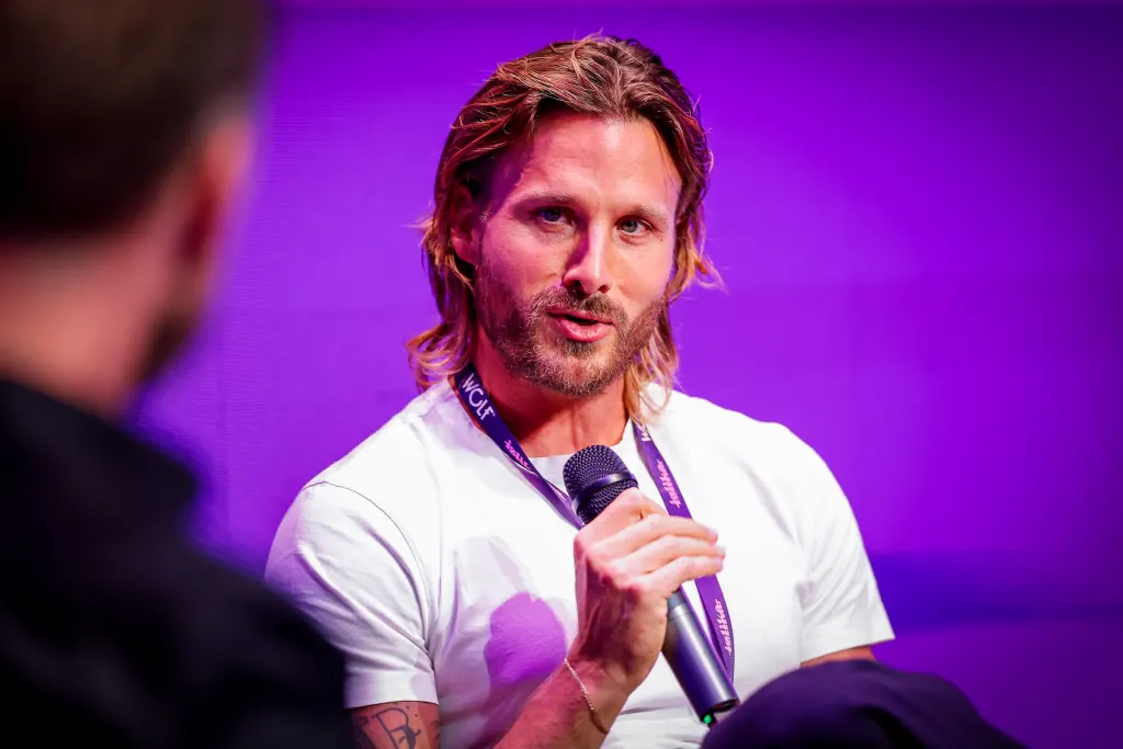 A person with long hair and a beard speaks into a microphone while wearing a lanyard, set against a vibrant purple background.