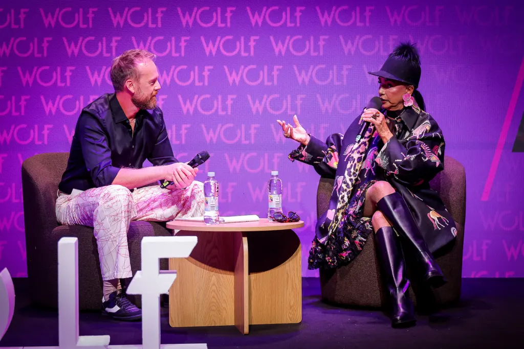 Two individuals engage in a conversation on stage, seated in armchairs with a small table between them, against a backdrop featuring repeated text.