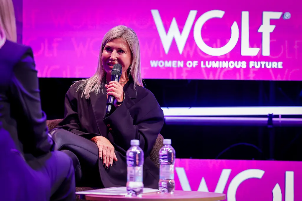 A person with long blonde hair speaks into a microphone on a stage at an event branded "WOLF Women of Luminous Future."