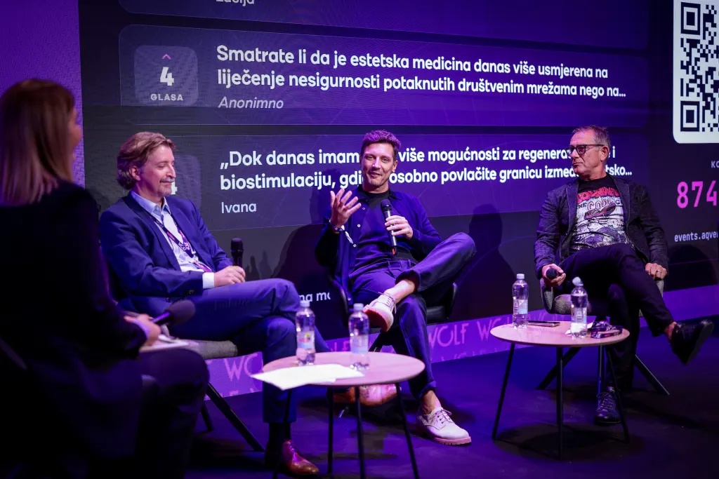 Three individuals engage in a discussion on stage during a conference, with a large screen displaying audience comments in the background.