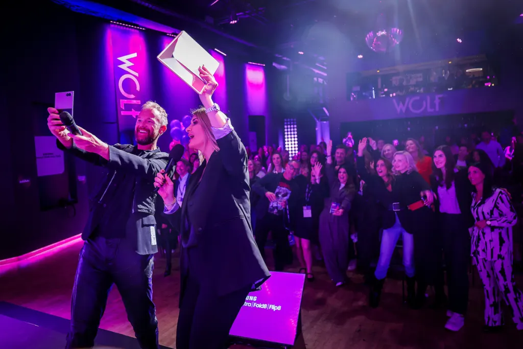 Two individuals on a stage at a WOLF event cheerfully pose for a selfie while holding a phone and a box, surrounded by an enthusiastic audience under vibrant purple lighting.