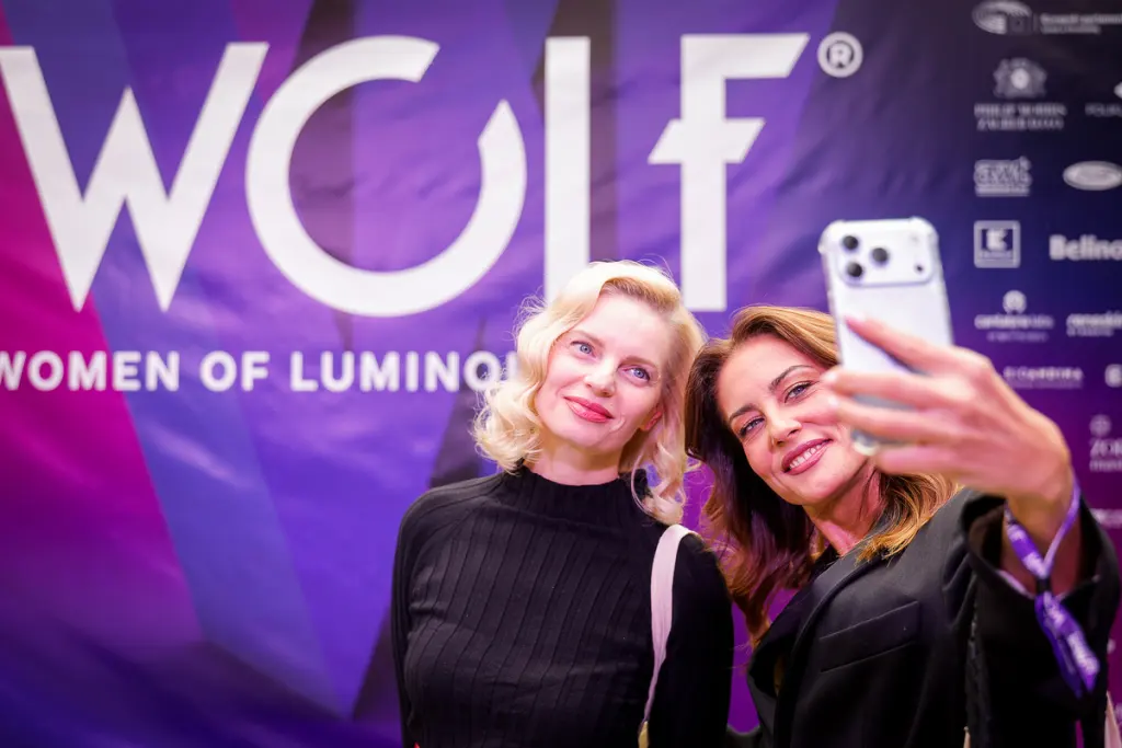Two individuals take a selfie in front of a purple backdrop featuring the text "WOLF WOMEN OF LUMINOUS".