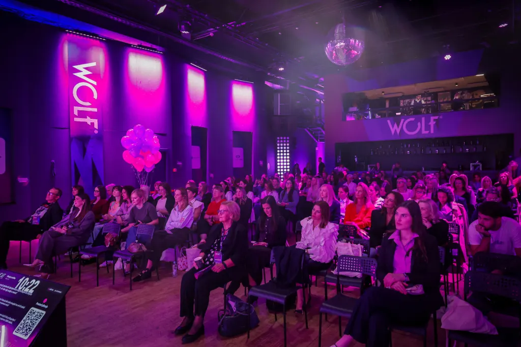A crowd of attendees sits in a dimly lit venue bathed in purple lighting, with "WOLF" branding visible on the walls and a disco ball hanging overhead.