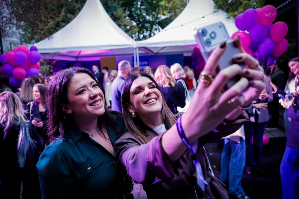 Two individuals take a selfie together at an outdoor event under purple lighting, surrounded by balloons and other attendees.