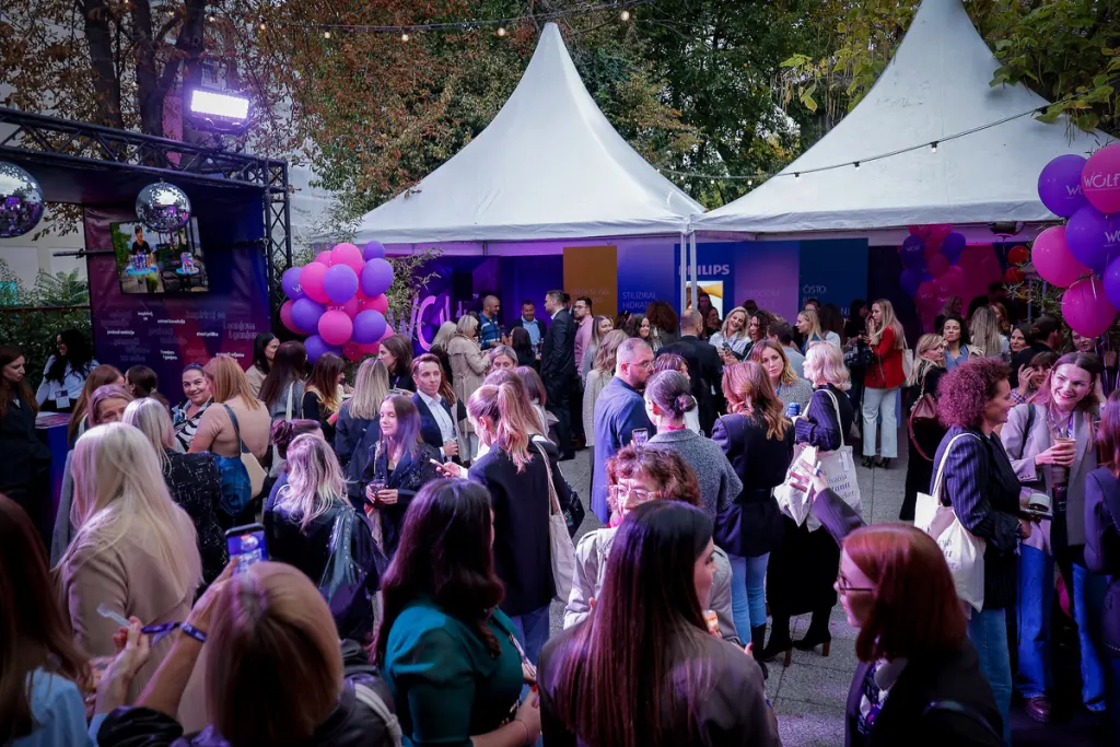 A lively outdoor gathering with people mingling under white tents, surrounded by purple and pink balloons and festive lighting.